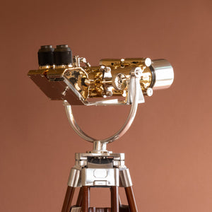 Close up of side view of Krauss of France brass and aluminium binoculars with the eye pieces on the left and the lens on the right. The aluminium cradle and the top of the wood tripod. Against a orange pink background.