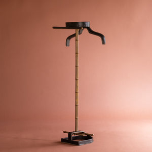 An angled side view of a 1950’s Valet de nuit; model 'Luxe' by Jacques Adnet, France. The vertical central tubular brass stand is made to look like bamboo. The base is leather in the shape of a horseshoe and it has a leather hanger at the top at an angle. 
