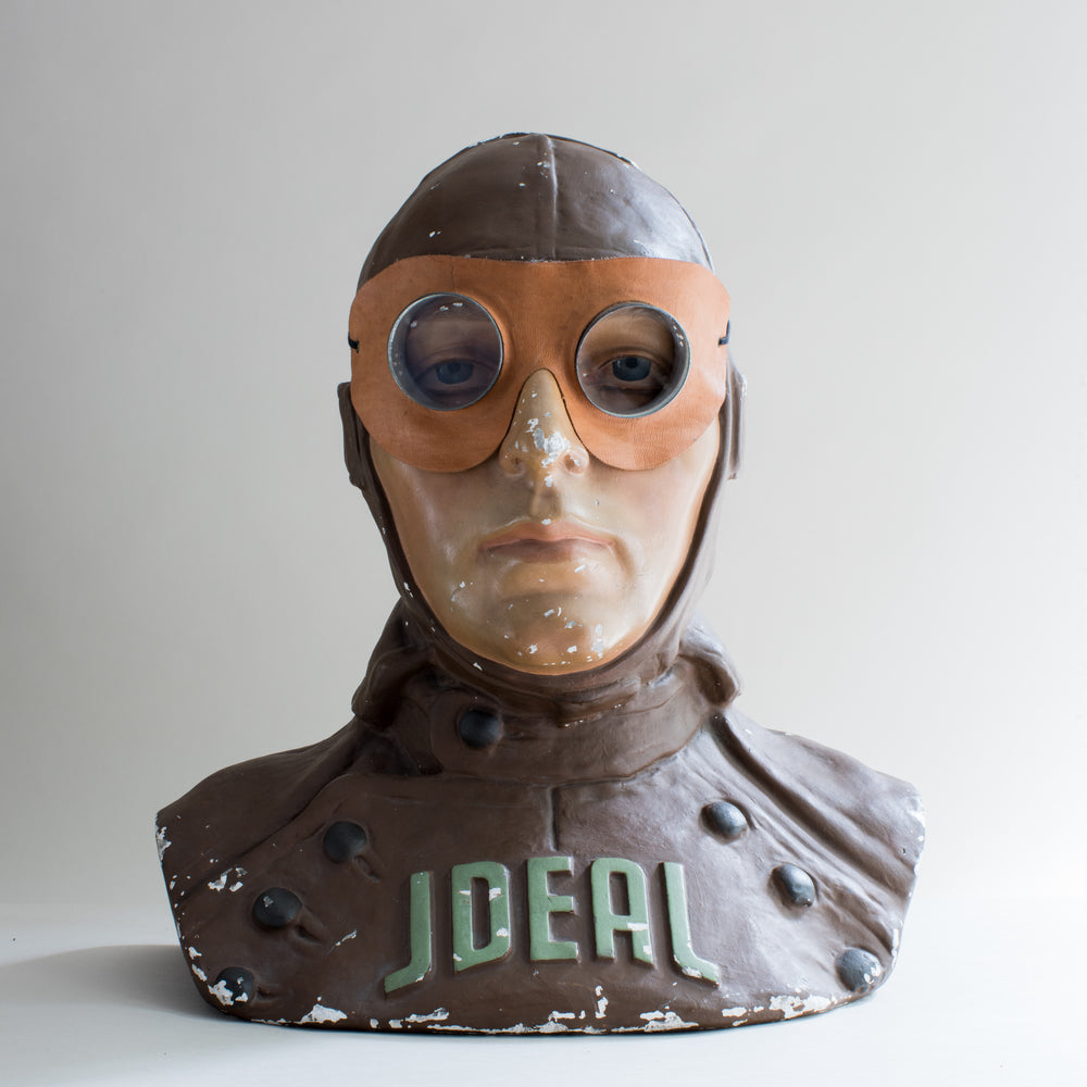 Front view of painted brown and beige plaster cast bust of air pilot wearing vintage motoring or aviation goggles with circular glass lens, metal disc surround and leather shaped mask with elastic strap by Kraus & Co. Word ideal across chest. White background.