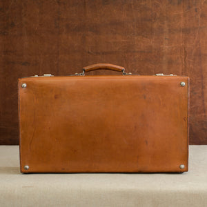 View of the bottom and corner studs of a English leather case, circa 1925, with handle and nickel plated brass locks at the top. Brown and cream background.