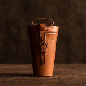 The image shows the original tan leather cover of a set of six horn beakers, circa 1880. The leather cover is showing the strap and leather covered buckle at the front of the case and a carrying leather strap at the top of the case. It is sitting on a brown background.