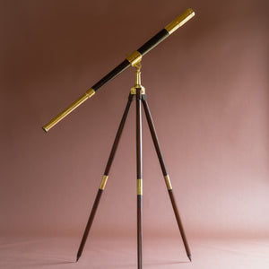 Side view of a leather covered brass telescope by J.J. Hicks dated 1901. Shown at a horizontal angle on a wooden and brass campaign tripod stand, by W. Watson & Sons London dated 1888. The telescope eyepiece is on the mid left and on the top right side is the end with the lens cap, positioned on top of brass and wood splayed tripod legs. The background is pink.