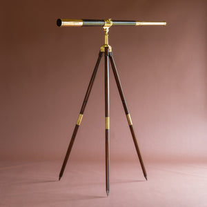 Side view of a leather covered brass telescope by J.J. Hicks dated 1901. Shown at a horizontal angle on a wooden and brass campaign tripod stand, by W. Watson & Sons London dated 1888. The telescope eyepiece is on the top right and the top left side is the end with the lens, positioned on top of brass and wood splayed tripod legs. The background is pink.