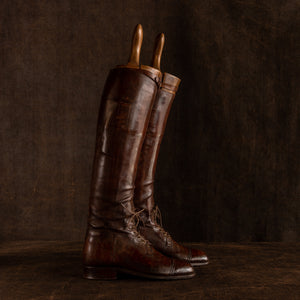 Side view of a pair of brown leather polo boots including their wooden trees made for Earl Spencer, Althorp Estate. They are laced up and placed sideways, rear boot slightly in front on a brown background.