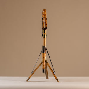 The image shows an Edwardian shooting stick with the tripod base open and the seat at the top closed together. Made of beech wood, brass and steel, circa 1910. The stool sits central on a beige and cream background with tripod legs splayed and the seat closed facing forwards.