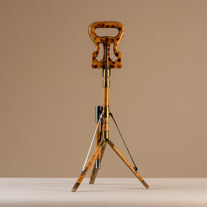 The image shows an Edwardian shooting stick with the tripod base open and the seat at the top closed together. Made of beech wood, brass and steel, circa 1910. The stool sits central on a beige and cream background with tripod legs splayed and the seat closed facing sideways.