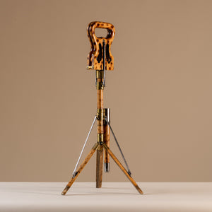 The image shows an Edwardian shooting stick with the tripod base open and the seat at the top closed together. Made of beech wood, brass and steel, circa 1910. The stool sits central on a beige and cream background with tripod legs splayed and the seat closed facing a diagonal angle.