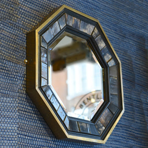 View from a diagonal of the below left side and front of a 1970’s Antony Redmile octagonal brass framed bevelled glass mirror with inset horn and ebonised wood surround with a blue woven background.