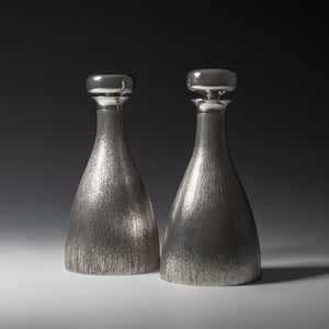 Front view of two silver 'bark finish' decanter with polished silver stoppers side by side, with hallmarks, London 1970’s with makers mark HOL - House of Lawrian, against a grey background.