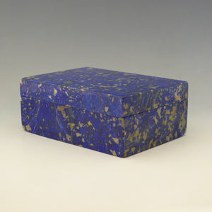 Front view of a box made from Lapis Lazuli at an angle with front right corner in the foreground. It is a cobalt blue base colour with beige and dark blue mottling running through the stone. Makers mark for George Betjemann and Sons, 1928. White background.