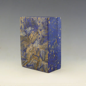 View of the base and front of a box made from Lapis Lazuli on it’s side. It is a cobalt blue base colour with beige and dark blue mottling running through the stone. The base has the angular striated lines of the stone. Makers mark for George Betjemann and Sons, 1928. White background.