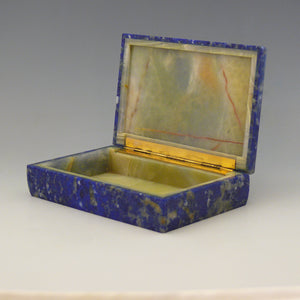 A front view at an angle of an open stone box with a yellow stone inside and gilt hinge along the edge adjoining the lid. The outside of the box is Lapis Lazuli with a cobalt blue base colour and white, beige and dark blue mottling running through the stone. white background.