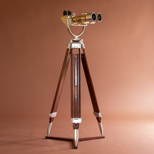 Full length side view of Krauss of France brass and aluminium binoculars on a wood tripod with lens facing right on a diagonal and the eye pieces to the left side. Against a orange pink background.