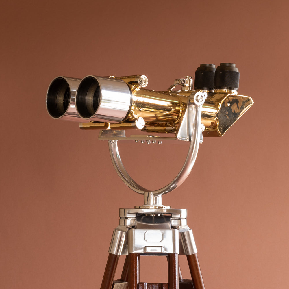 Side view of Krauss of France brass and aluminium binoculars with eye pieces on the right side, lens to the left side. Bottom view of top of wood tripod stand with deep pink background.