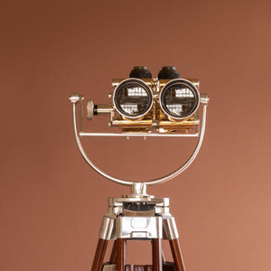Close up of front view of Krauss of France brass and aluminium binoculars with the lens at the front centre. The aluminium cradle and the top of the wood tripod. Against a orange pink background.