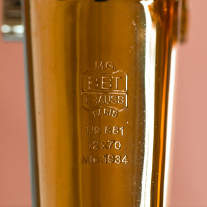Close up of the engraved manufacturers stamp into the brass of the Krauss of France binoculars  Against a orange pink background.