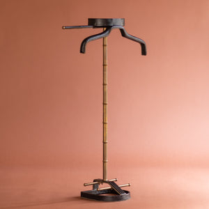 An angled front view of a 1950’s Valet de nuit; model 'Luxe' by Jacques Adnet, France. The vertical central tubular brass stand is made to look like bamboo. The base is leather in the shape of a horseshoe and it has a leather hanger at the top at an angle. 