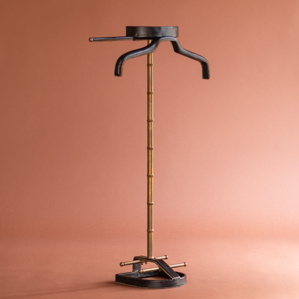 An angled front view of a 1950’s Valet de nuit; model 'Luxe' by Jacques Adnet, France. The vertical central tubular brass stand is made to look like bamboo. The base is leather in the shape of a horseshoe and it has a leather hanger at the top at an angle. 