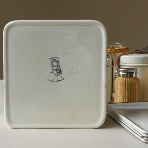 Close up of items from the motoring picnic case made by G. W. Scott and Sons trading under their brand name ‘Coracle’ ; circa 1920. Showing enamelled metal food boxes with plated tops with maker’s stamp, wicker covered glass flasks, ceramic butter and preserve pots, plated tea/sugar tin, four enamelled metal plates and one of four ceramic cups & saucers. Beige and white background.
