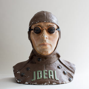 Front view of painted brown and beige plaster cast bust of air pilot wearing vintage motoring or aviation goggles with fine mesh and fabric strap by Kraus & Co, facing front. Word ideal across chest. White background.