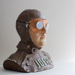 Side view of painted brown and beige plaster cast bust of air pilot wearing vintage motoring or aviation goggles with circular glass lens, metal disc surround and leather shaped mask with elastic strap by Kraus & Co. Word ideal across chest. White background.