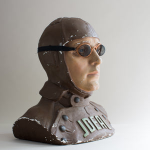 Side view of painted brown and beige plaster cast bust of air pilot wearing vintage motoring or aviation goggles with fine mesh and fabric strap by Kraus & Co, facing right. White background.
