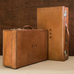 A matching pair of English leather cases, circa 1925. On the left the suitcase is at a diagonal angle with the embossed initials F.I.W. lid facing the right and handle at the top. On the right the suitcase initialed  R.E.W. is on it’s side with the handle facing right. Both have nickel plated brass locks. Brown and cream background.