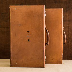 View of the tops of a matching pair of English leather cases, circa 1925, one in front of the other, handles facing the right side. The left one has the initials  F.I.W., the right suitcase sits slightly behind. The  nickel plated brass locks just show on the right sides of the suitcases. Brown and cream background.