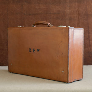 View at a diagonal angle of the lid side of the leather case, circa 1925, handle at the top, just showing nickel plated brass locks at the top and the initials R.E.W. Brown and cream background.