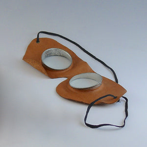 Vintage motoring or aviation goggles with circular glass lens, metal disc surround and leather shaped mask with elastic strap by Kraus & Co viewed on an angle. White background.