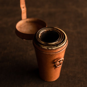 The image is a view from above the opened original tan leather cover of a set of six horn beakers, circa 1880. The leather cover is opened with the leather strap unfastened, showing the leather covered buckle and the tops of the six horn beakers. The opened tan leather lid is showing against the brown background.