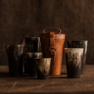 The image shows the original closed tan leather case sitting with six horn beakers, showing the strap and leather covered buckle at the front of the case and a carrying leather strap at the top of the case. They are sitting on a brown background, the leather case is sitting right of centre with four horn beaker of varying sizes on the left side and two different sized horn beakers on the right side.