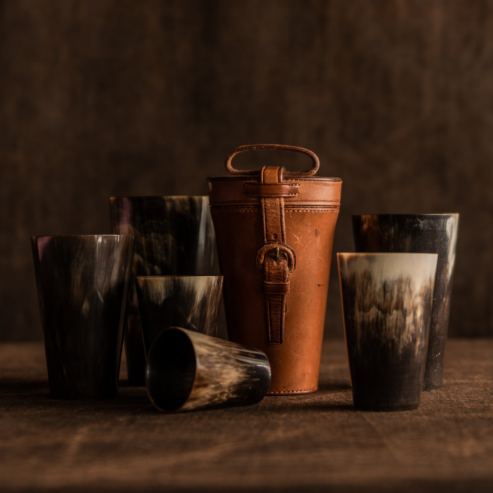 The image shows the original closed tan leather case sitting with six horn beakers, showing the strap and leather covered buckle at the front of the case and a carrying leather strap at the top of the case. They are sitting on a brown background, the leather case is sitting right of centre with four beakers on horn beaker of varying sizes on the left side, one lying on it’s side and two different sized horn beakers on the right side.