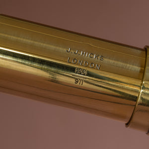 Close up of a leather covered  brass telescope by J.J. Hicks dated 1901, showing the maker’s name and date engraved into the brass. Against a pink background.