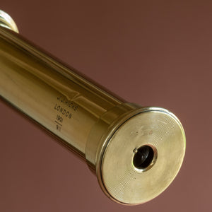 Close up of a leather covered brass telescope by J.J. Hicks dated 1901. Showing the engraving details of the maker’s name and date stamp and the integral swivel dust cover in the eyepiece. Against a pink background.