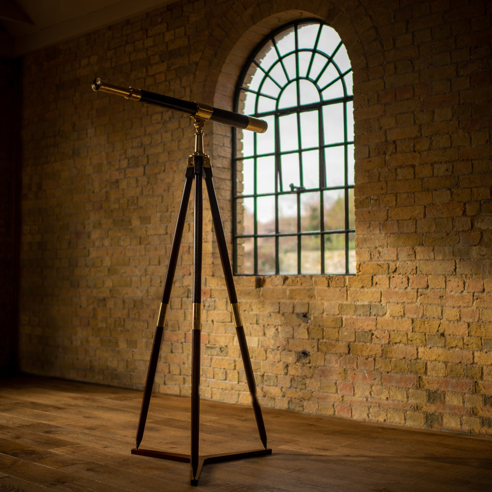 Side view of a leather covered brass telescope by J.J. Hicks dated 1901. Shown at an angle on a wooden and brass campaign tripod stand, on a wooden floor pointing out towards an arched window set into a brick wall, the the brass eyepiece to the left side, the window on the right.