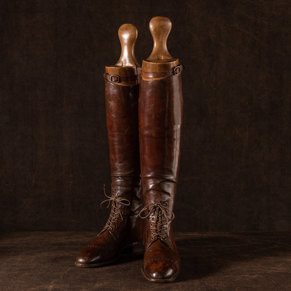 Front view of a pair of brown leather polo boots including their wooden trees made for Earl Spencer, Althorp Estate. They are laced up and at a three quarter position on a brown background.