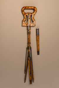 The image is from above, it shows an Edwardian shooting stick with the tripod base and the seat at the top closed together. To the right of shooting stick is the cover for the tripod legs to make the seat into a shooting stick. All made of beech wood, brass and steel, circa 1910. They are against a beige and cream background.