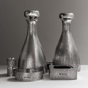 Front view of two silver 'bark finish' decanter with polished silver stoppers side by side, with hallmarks, London 1970’s with makers mark HOL - House of Lawrian, with silver ashtray to front right, silver coaster front left, silver spirit measure left side. Against a grey background.
