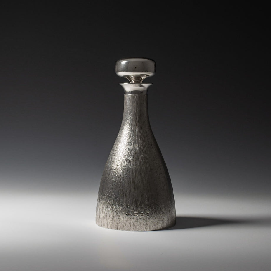 Front view of silver 'bark finish' decanter with polished silver stopper with hallmark, London 1973 with makers mark HOL - House of Lawrian, against a grey background.