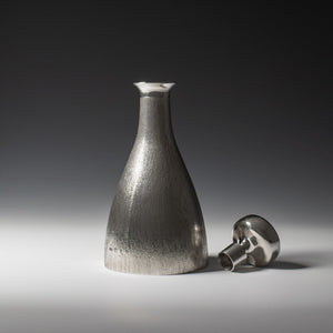 Front view of silver 'bark finish' decanter with polished silver stopper to the right of the picture with hallmark, London 1973 with makers mark HOL - House of Lawrian, against a grey background.