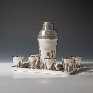 Walker and Hall silver plated rectangular tray with raised edges viewed from the side with a handle showing right hand side. Silver cocktail shaker on centre of tray, with three silver cocktail beakers either side of tray. White and grey set at a diagonal.
