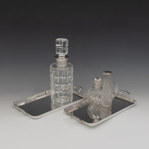 View of top of a pair of Walker and Hall silver plated rectangular trays with raised edges with a handle each end. The left tray has a cut glass decanter with silver collar on it. The right one has a wrythen glass and silver topped perfume bottle and to the right of it a silver topped wrythen glass jug. White and grey background set at a diagonal.