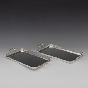 View of top of a pair of Walker and Hall silver plated rectangular trays with raised edges with a handle each end. White and grey background set at a diagonal to the left side.