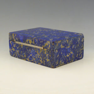 Rear view of a box made from Lapis Lazuli at an angle with rear left corner in the foreground. The gilt hinge runs across the length of the back. It is a cobalt blue base colour with beige and dark blue mottling running through the stone. Makers mark for George Betjemann and Sons, 1928. White background.