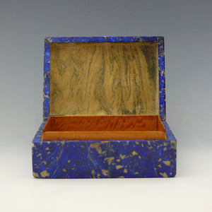 Front view of a box with the lid open, made from Lapis Lazuli with a cobalt blue base colour and beige and dark blue mottling running through the stone. Inside the base is lined in satinwood, the lid is a yellow cream coloured stone flecked with grey. Makers mark for  George Betjemann and Sons, London 1928. White background.