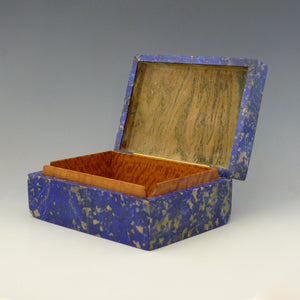Front view from an angle of a box with the lid open, made from Lapis Lazuli with a cobalt blue base colour and beige and dark blue mottling running through the stone. Inside the base is lined in satinwood, the lid is a yellow cream coloured stone flecked with grey. Makers mark for  George Betjemann and Sons, London 1928. White background.