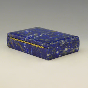 A view from an angle with the back left corner in the foreground of a Lapis Lazuli box with a cobalt blue base colour and white, beige and dark blue mottling running through the stone and a gilt hinge along the rear edge. White background. 