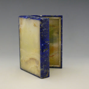 An open stone box resting on it’s side with the lid open viewed from below. The base and inside are yellow stone. The outside of the box is Lapis Lazuli with a cobalt blue base colour and white, beige and dark blue mottling running through the stone. white background.