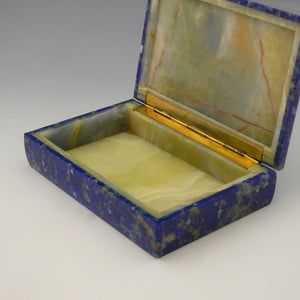 Detail of the front view at an angle of an open stone box with a yellow stone inside and gilt hinge along the edge adjoining the lid. The outside of the box is Lapis Lazuli with a cobalt blue base colour and white, beige and dark blue mottling running through the stone. white background.